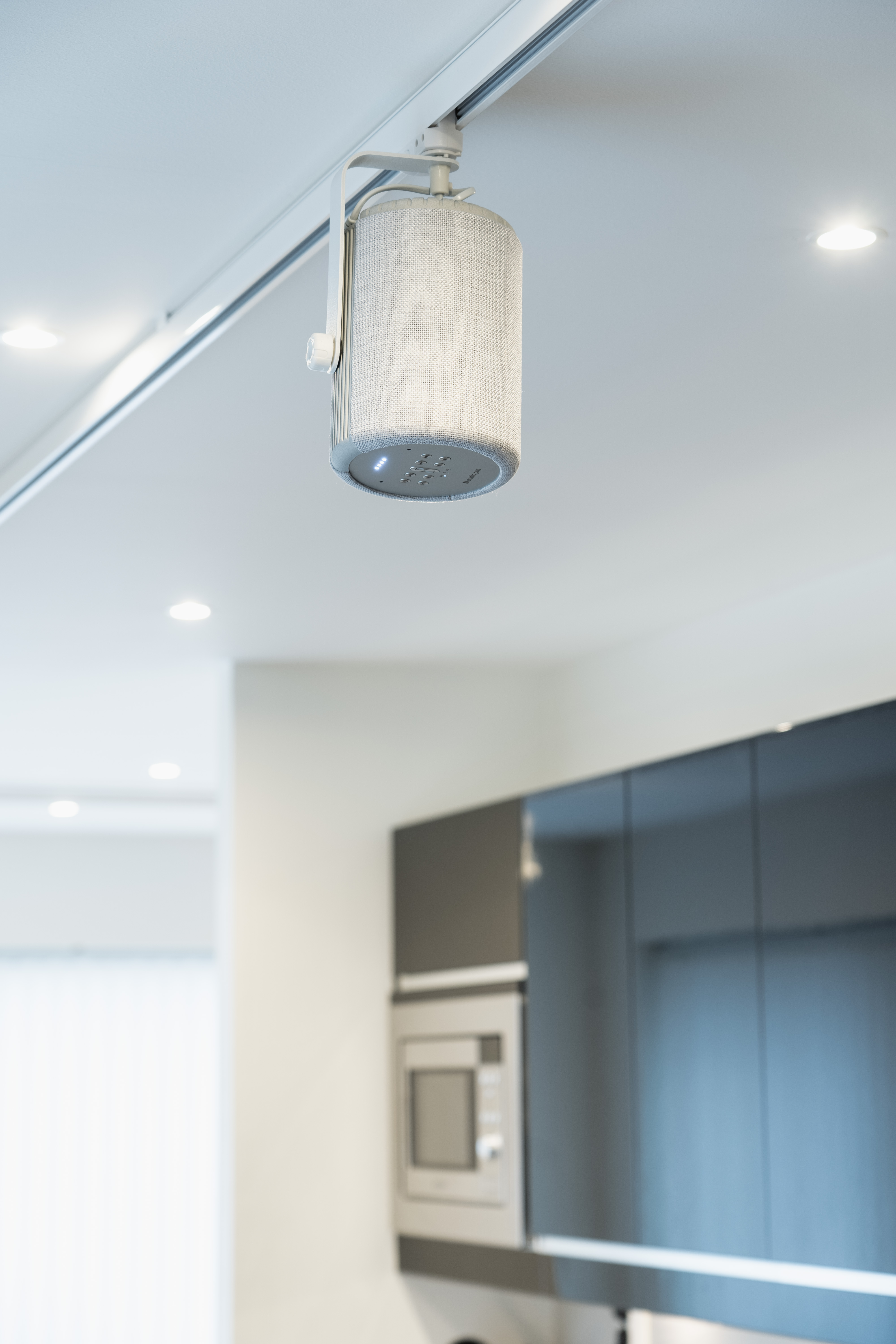 Biały głośnik sufitowy wisi w kuchni. Tło to czarne szafki i kuchenka mikrofalowa.