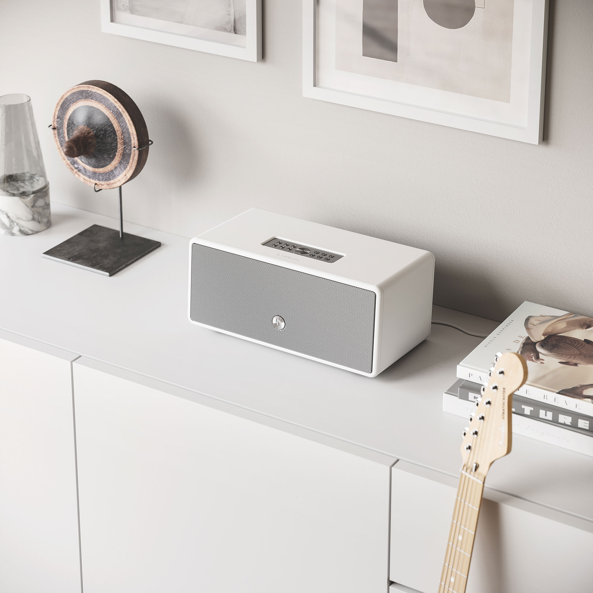 White speaker on a white surface with books and a guitar. Gray wall in background.