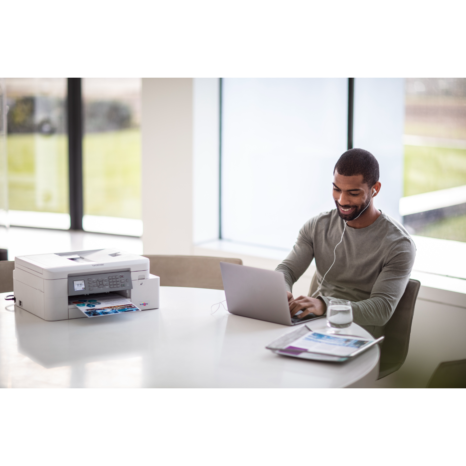 Mężczyzna pracujący przy okrągłym stole. Drukarka i laptop. Szklanka wody i magazyn.
