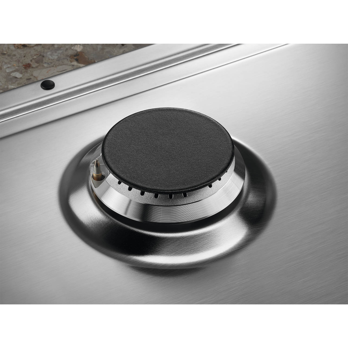 Zbliżenie palnika kuchenki gazowej. Czarna nasadka na srebrnej powierzchni. Blat granitowy w tle.