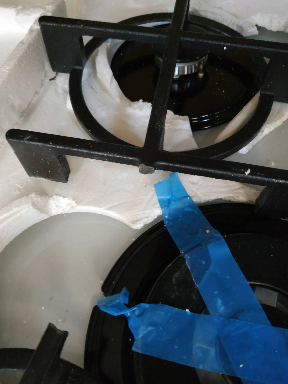 Zbliżenie na ruszty palników kuchenki gazowej i opakowanie ochronne, z niebieską taśmą.