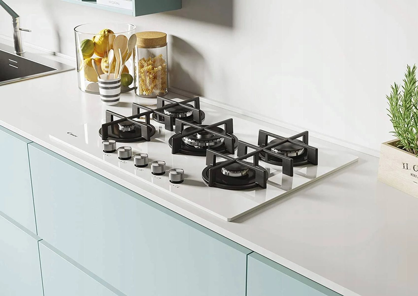 Biała kuchenka ze szklaną powierzchnią i srebrnymi pokrętłami, na jasnoniebieskiej szafce.