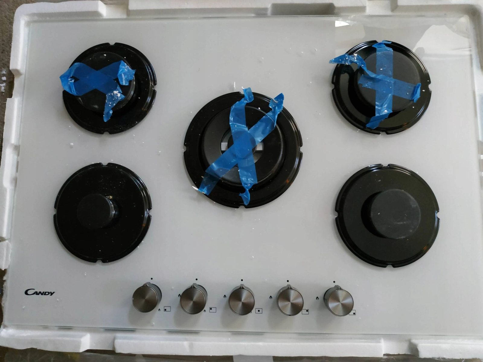 Biała płyta grzewcza Candy z pięcioma palnikami. Niebieska taśma zakrywa palniki. Srebrne pokrętła poniżej.