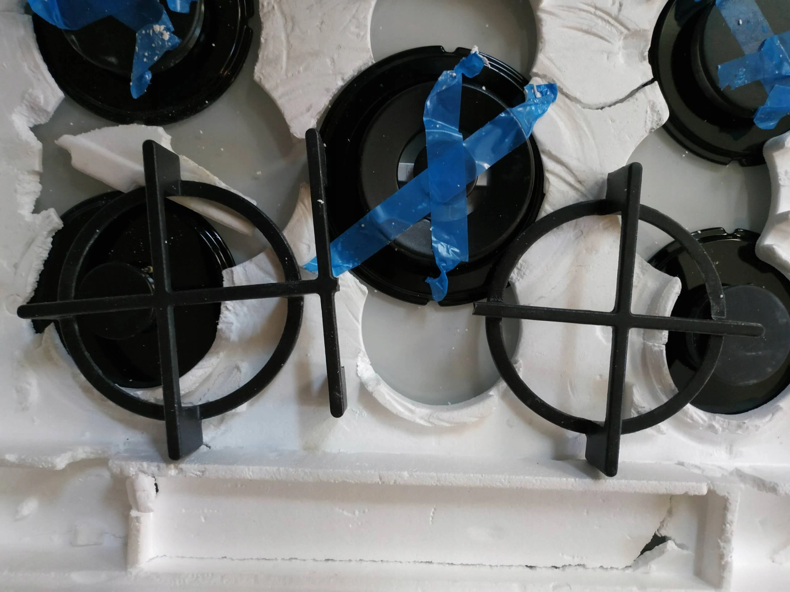 Zbliżenie na płytę gazową z czarnymi palnikami i pierścieniami podporowymi, biała powierzchnia.