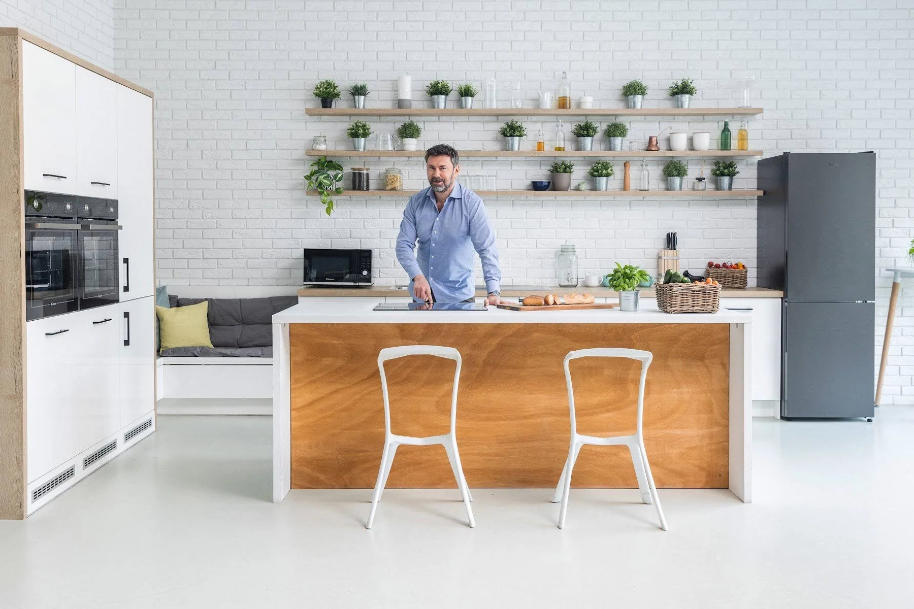 Mężczyzna w nowoczesnej kuchni. Białe szafki, wyspa i ceglana ściana. Rośliny na półkach. Dwa krzesła.