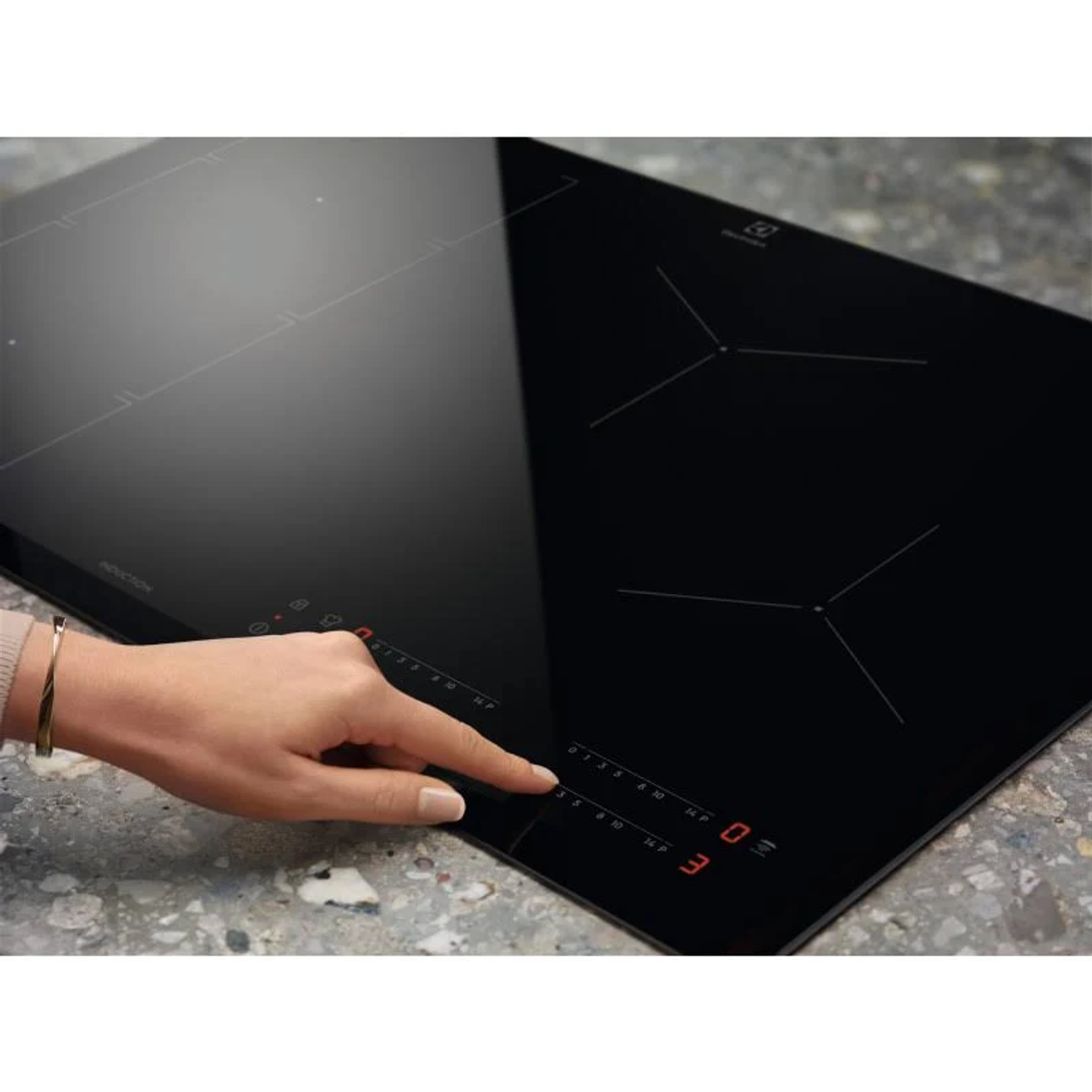 Dłoń dotyka czarnej płyty indukcyjnej, pokazując elementy sterujące i wyświetlacz cyfrowy. Widoczna marka Electrolux.