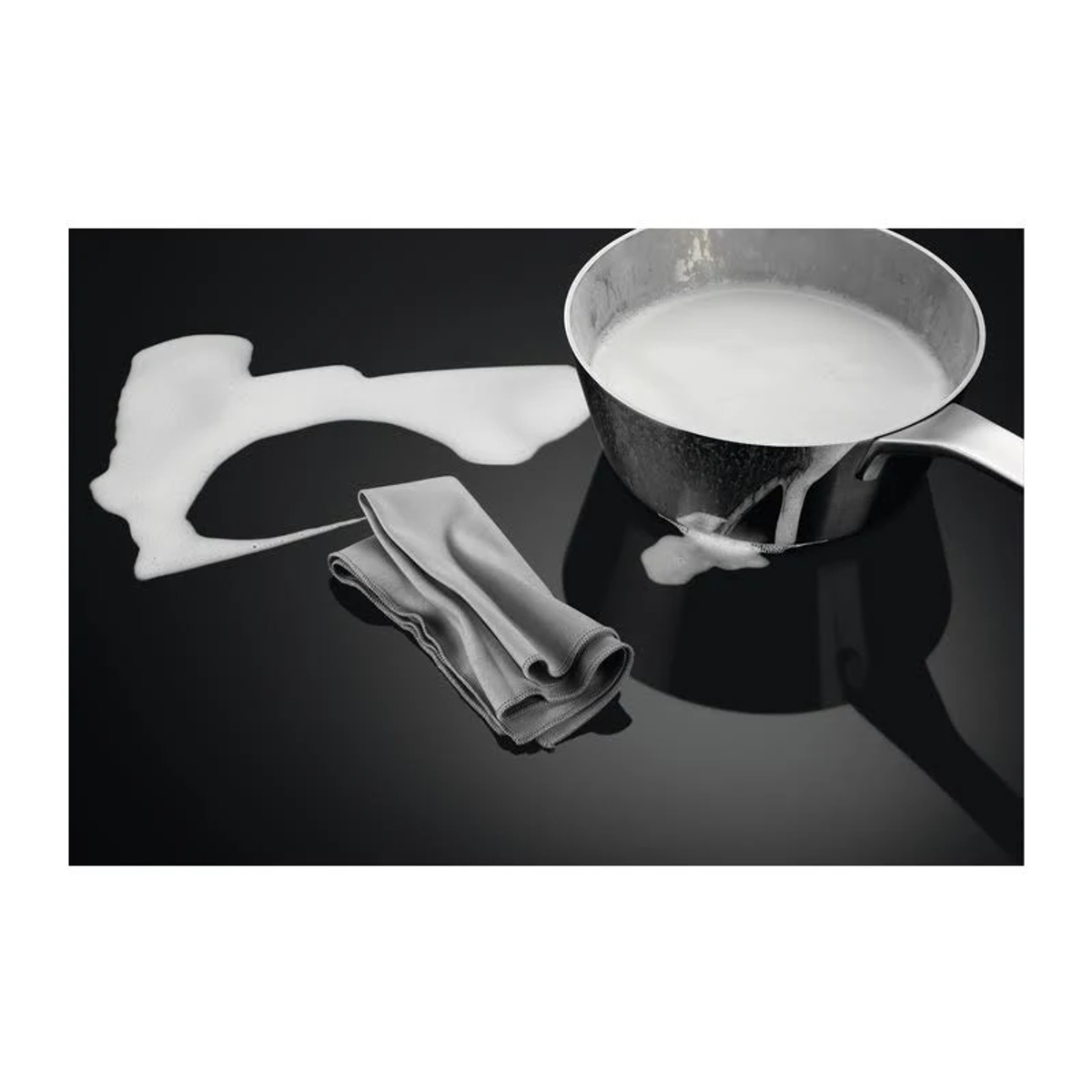 Garnek z wykipiałym mlekiem, szara ściereczka i czarna płyta kuchenna.