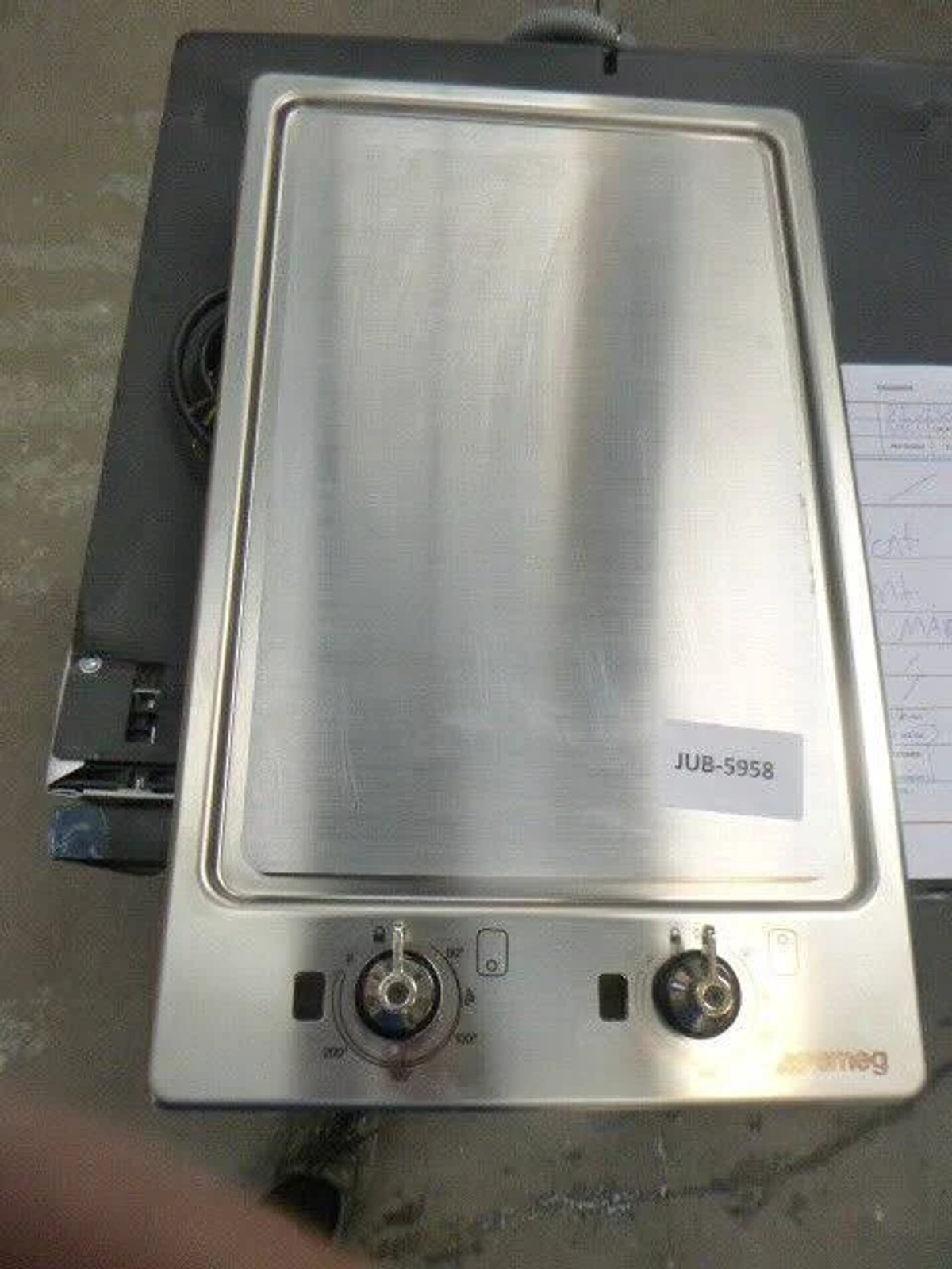 Un piano cottura Smeg in metallo con due manopole e un'etichetta. È sopra una superficie nera.