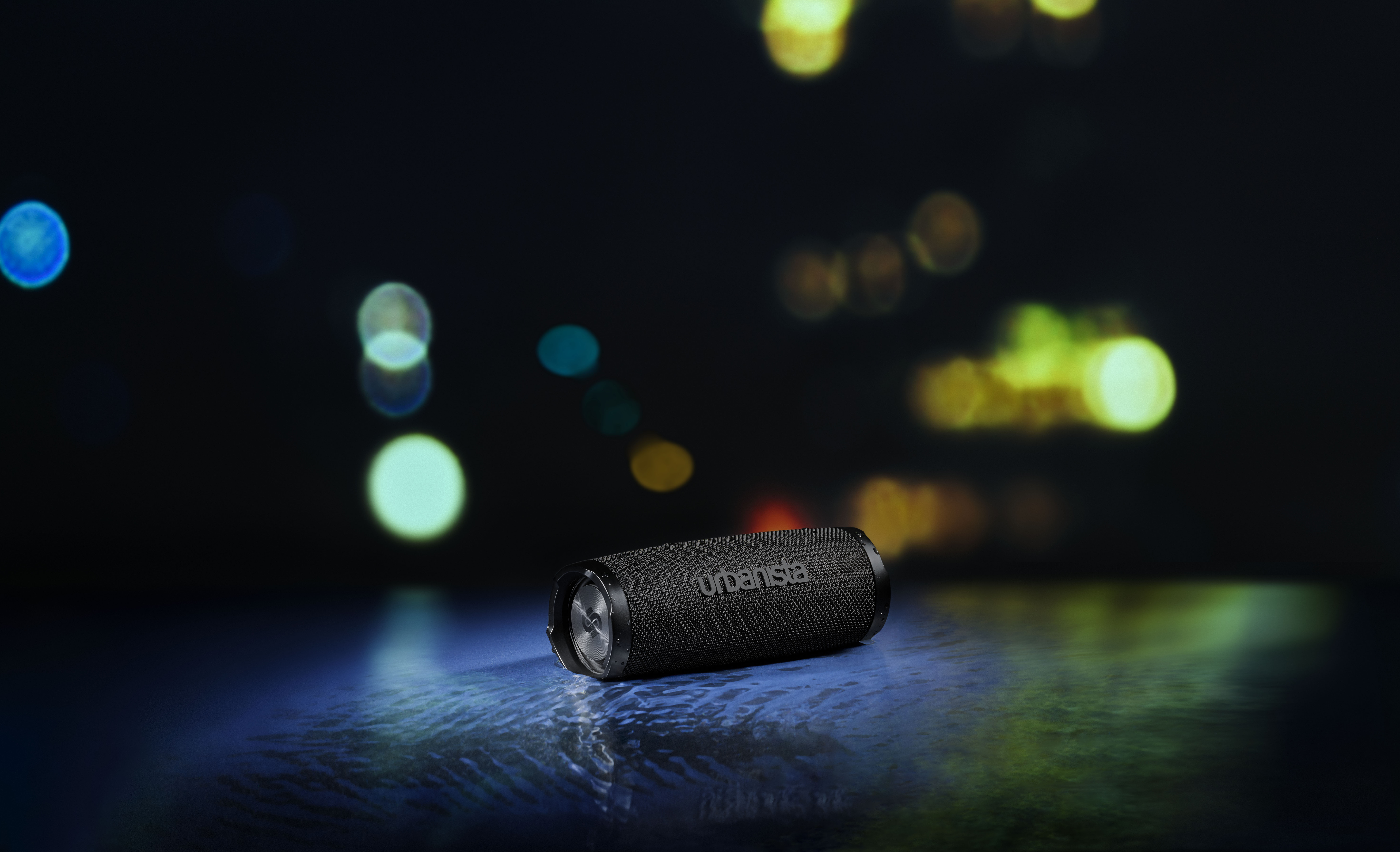 Czarny, cylindryczny głośnik na odblaskowej powierzchni, rozmyte tło miasta z światłami bokeh.