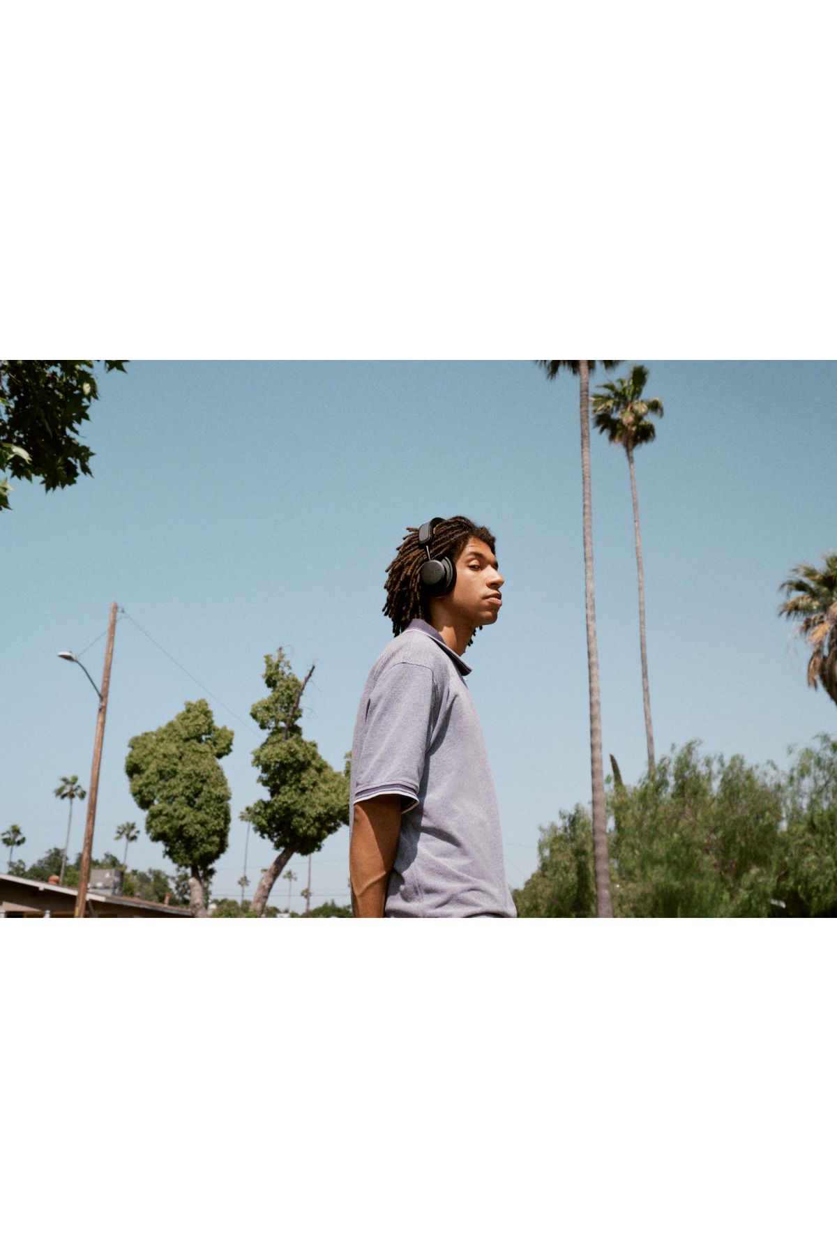 Młody mężczyzna z dredami w słuchawkach spaceruje na zewnątrz, widoczne są drzewa i błękitne niebo.