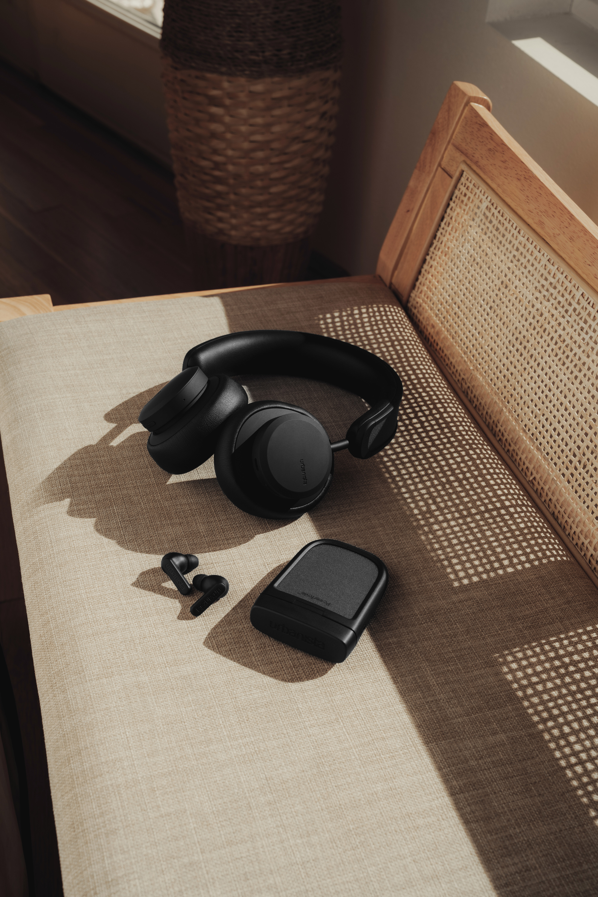 Czarne słuchawki, słuchawki douszne i etui ładujące na beżowym krześle. Ciepłe światło.