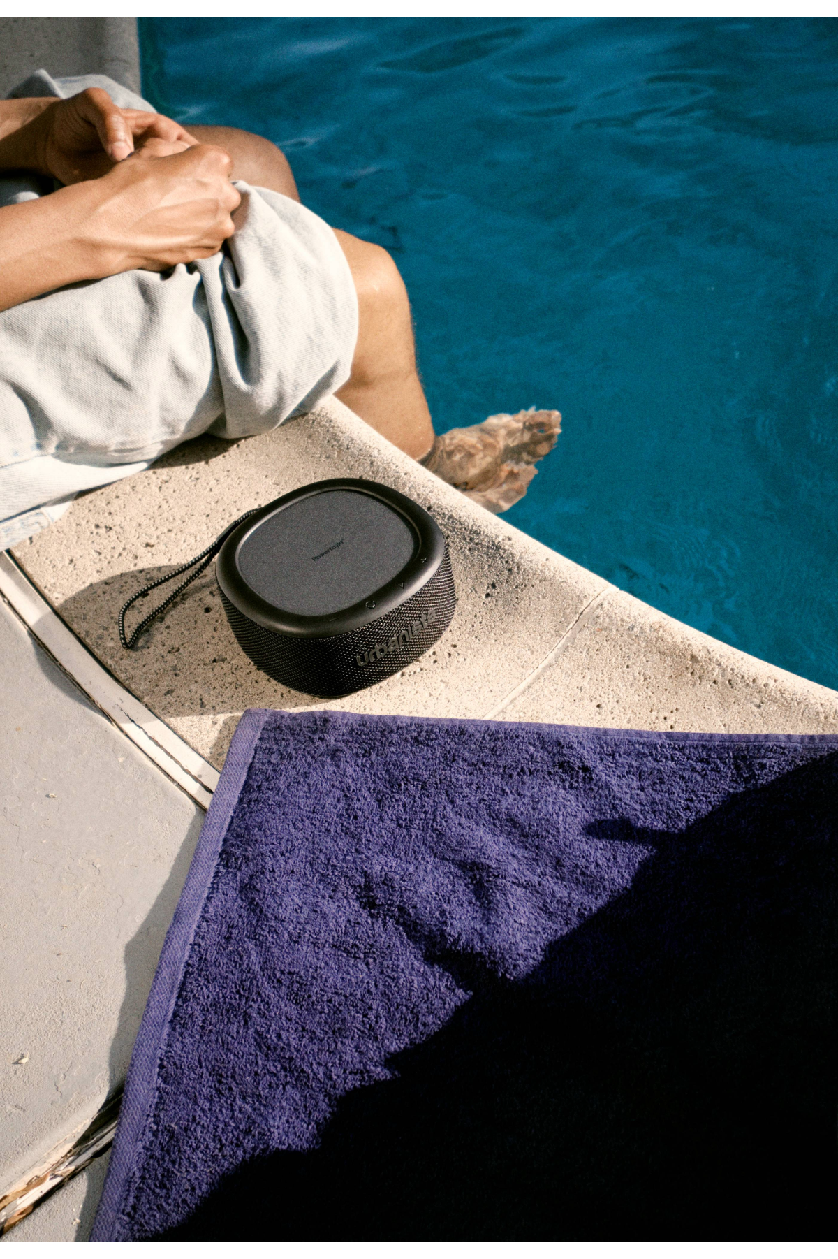 Haut-parleur noir sur rebord en pierre, jambes d'une personne, piscine bleue et serviette.