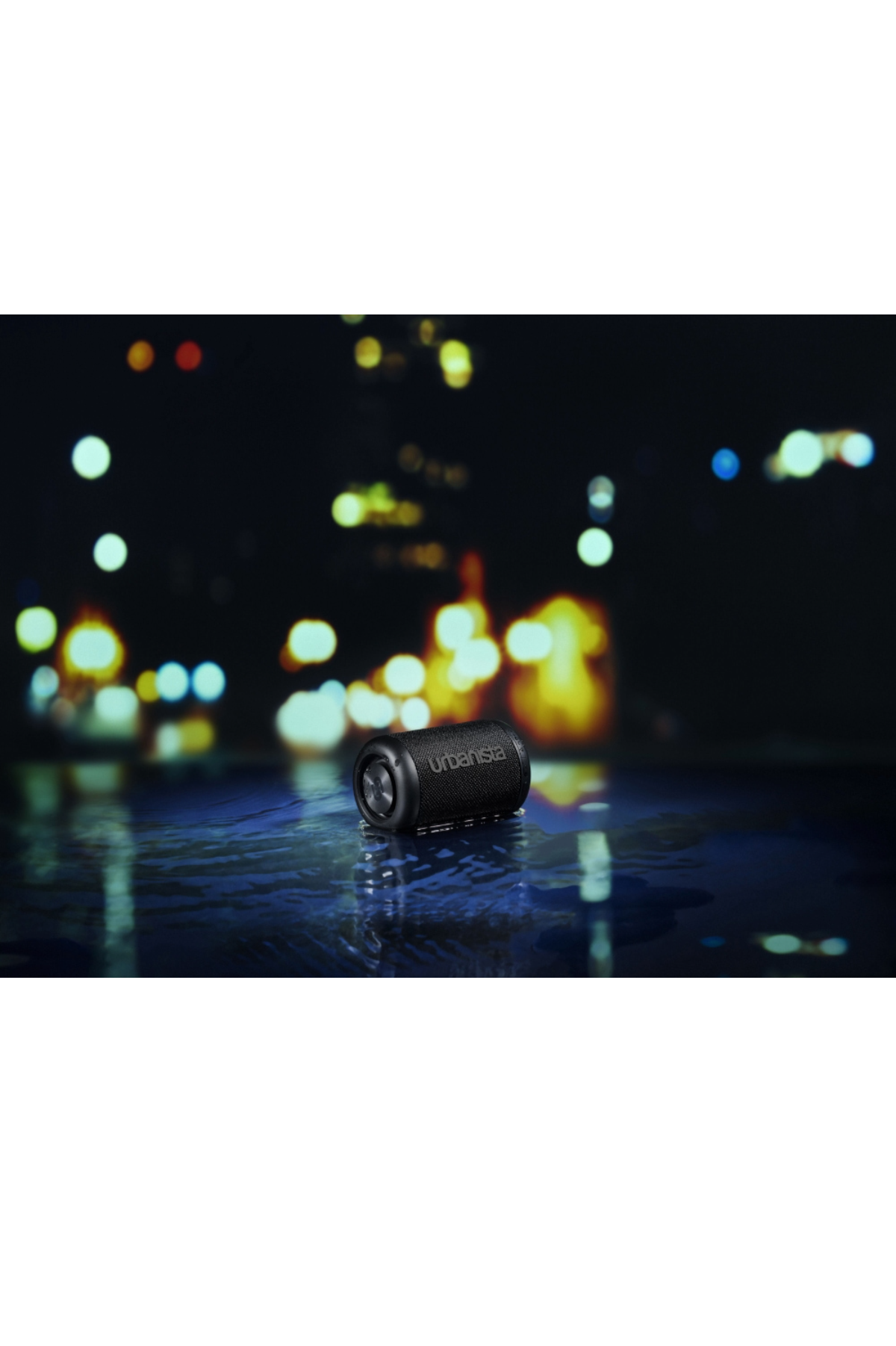 Czarny głośnik cylindryczny na mokrej powierzchni z rozmytymi światłami nocnymi.