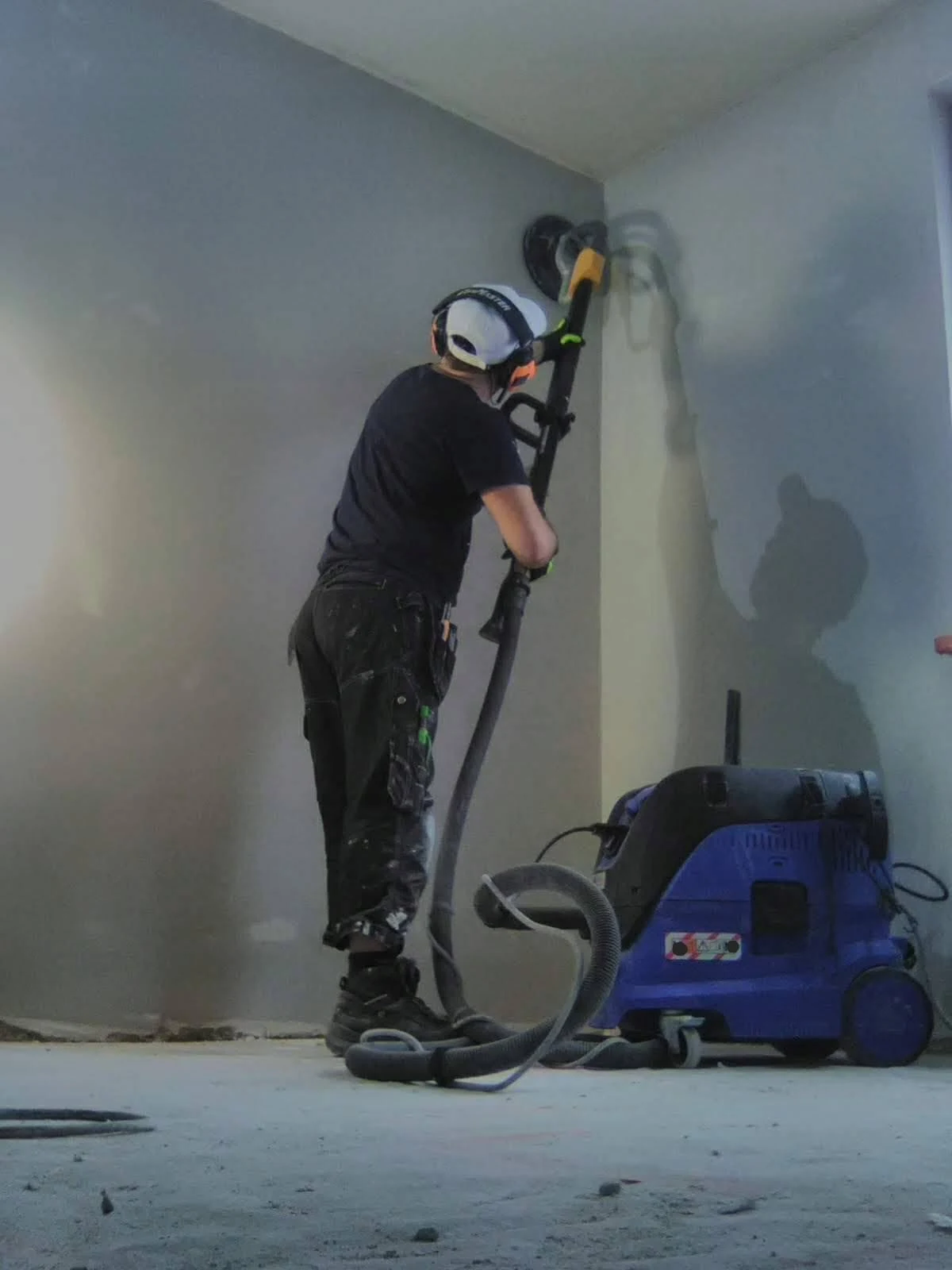 Una persona lijando una pared con una lijadora larga y una aspiradora.