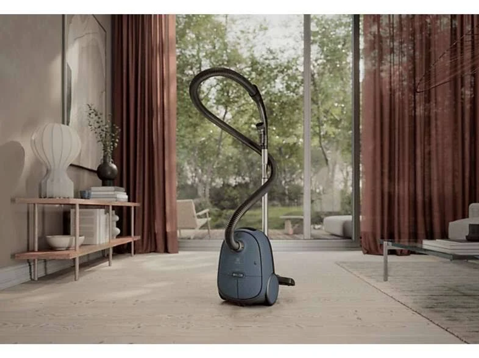 Niebieski odkurzacz stoi w pokoju z oknem i zasłonami, na drewnianej podłodze.