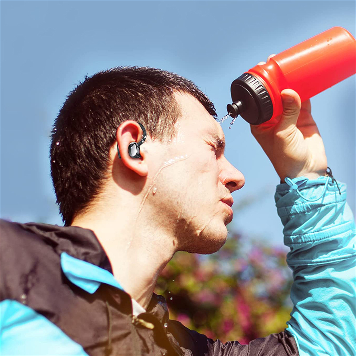 Ein Mann spritzt Wasser aus einer roten Wasserflasche in sein Gesicht. Er trägt einen Ohrhörer.