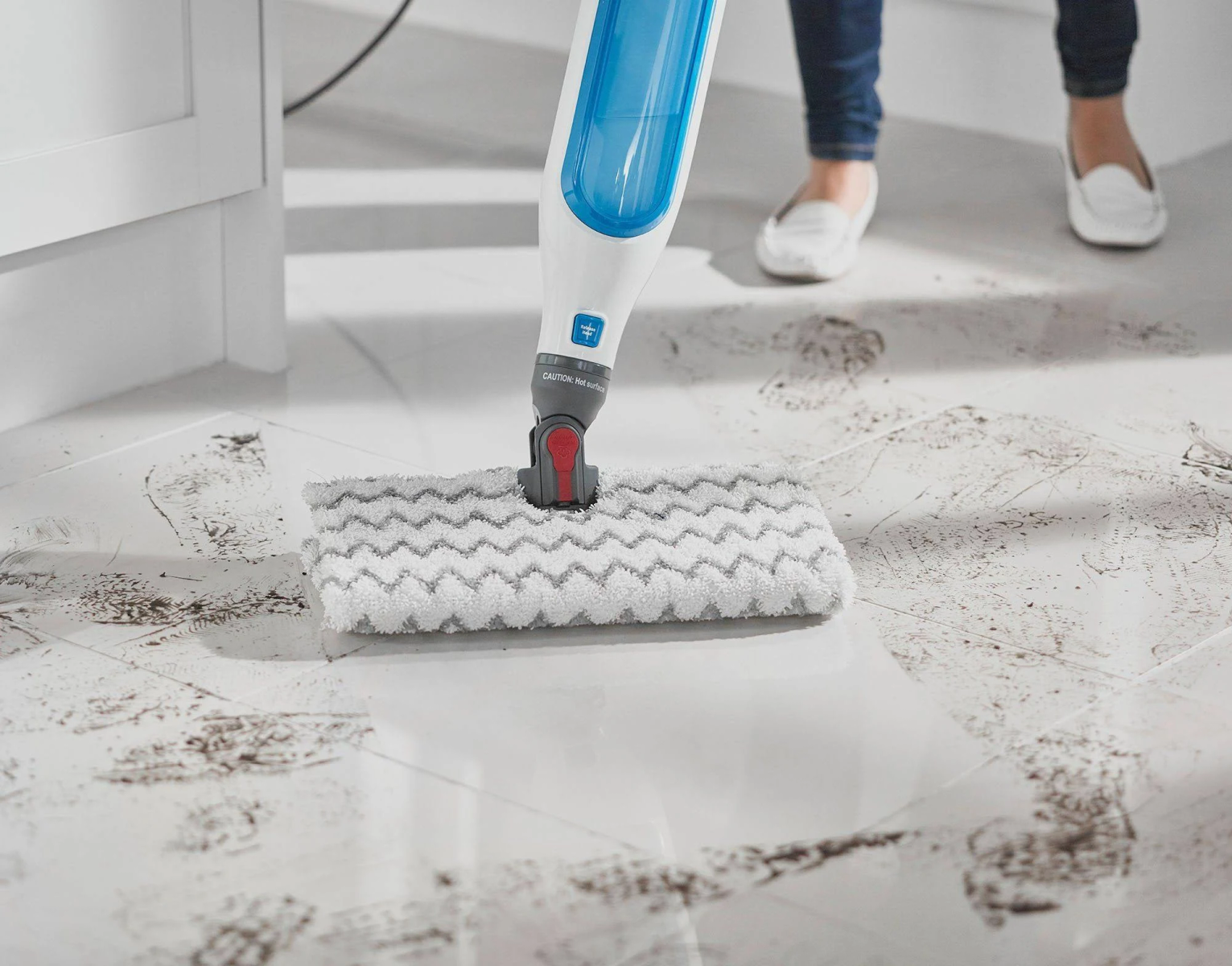 Osoba czyszcząca białą podłogę z płytek biało-niebieskim mopem parowym, pozostawiając mokre smugi i brudne ślady.