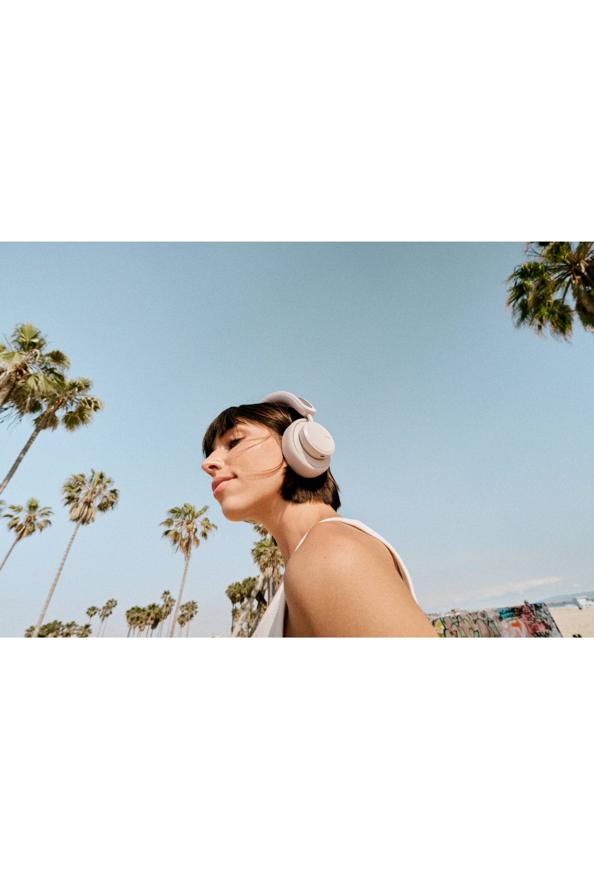Kobieta w słuchawkach, spoglądająca w górę, niebieskie niebo, palmy w tle.