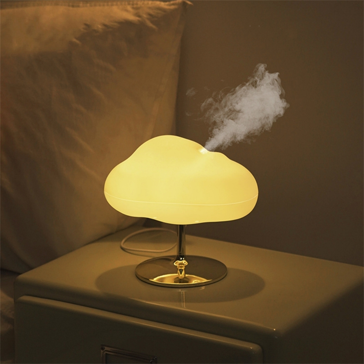 Eine wolkenförmige Lampe auf einem Nachttisch. Sie emittiert Dampf. Die Lampe ist weiß und hat einen goldenen Sockel.