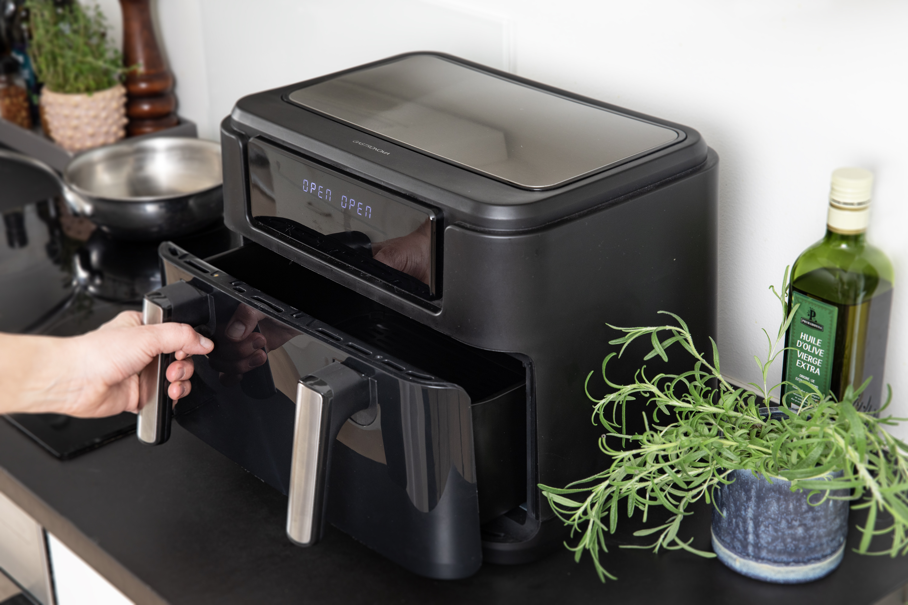 Czarna frytkownica z otwartą szufladą, dłoń trzymająca uchwyt, rozmaryn, oliwa z oliwek.