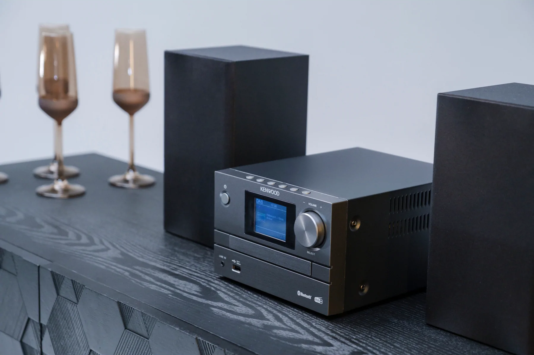 Equipo estéreo negro con altavoces, copas y mesa de madera. Fondo blanco.