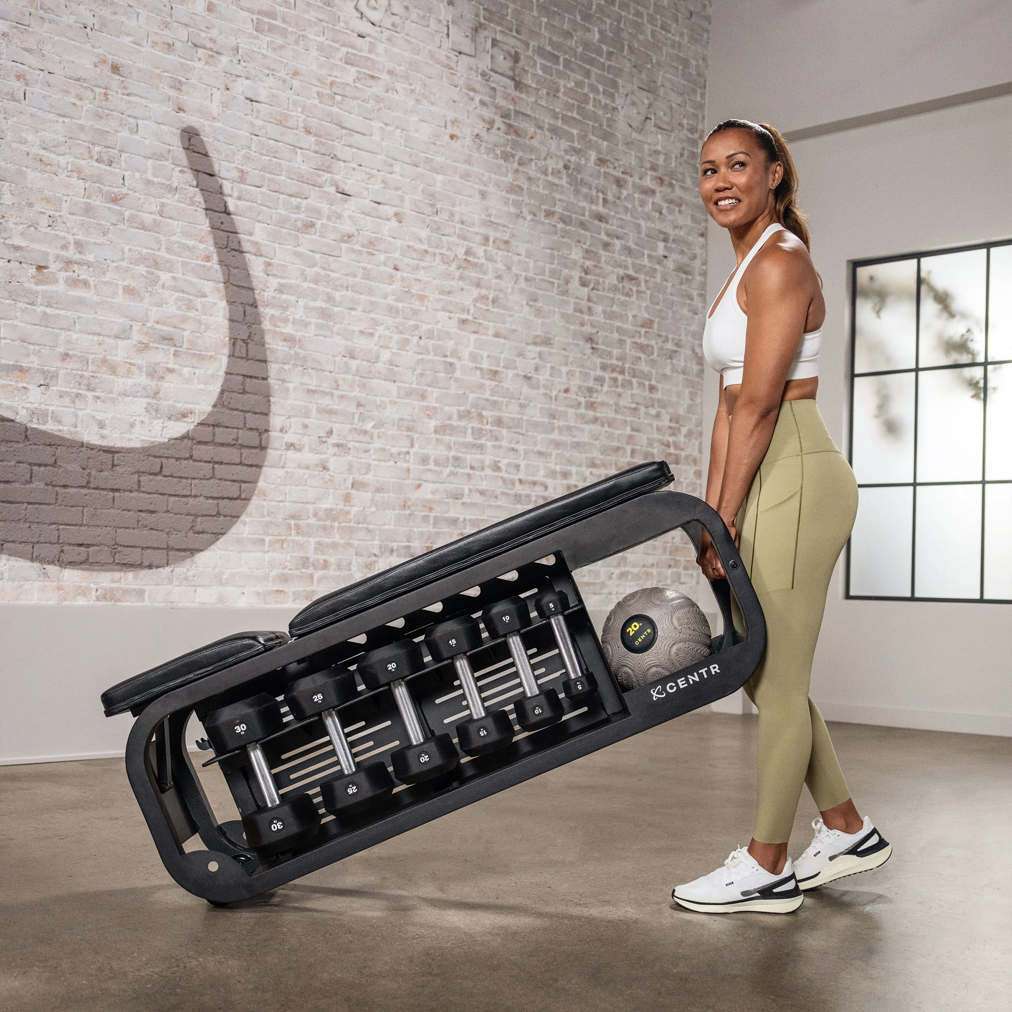 A woman holding a weight bench with dumbbells and a ball in a gym.