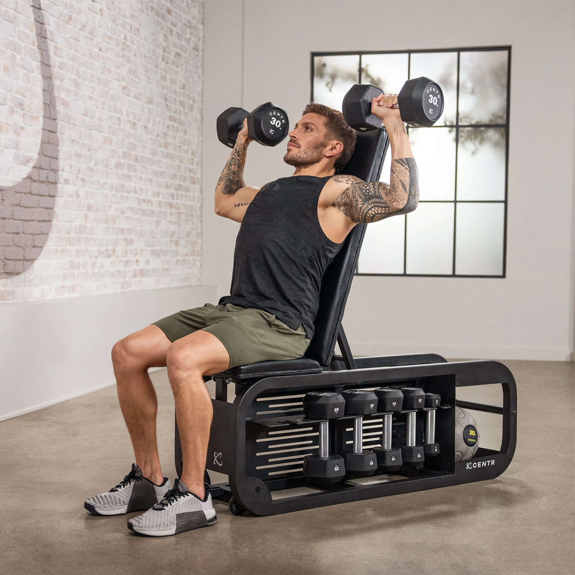 Man lifting dumbbells while seated on a weight bench, with other weights on the bench.
