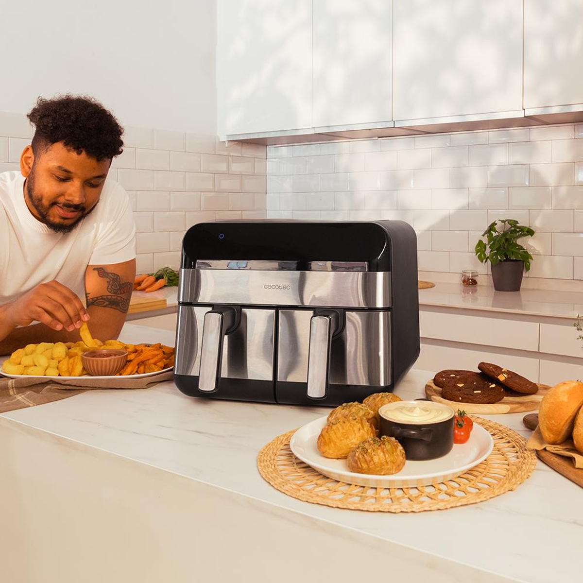 Osoba jedząca frytki i frytkownica powietrzna z podwójnym koszem z jedzeniem na blacie kuchennym, z innymi potrawami.