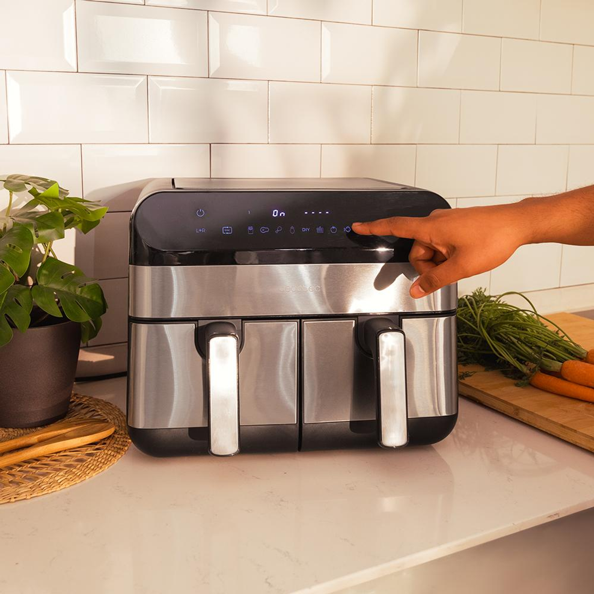 Frytownica powietrzna, czarna ze srebrnymi uchwytami, na blacie z ręką dotykającą panelu sterowania.