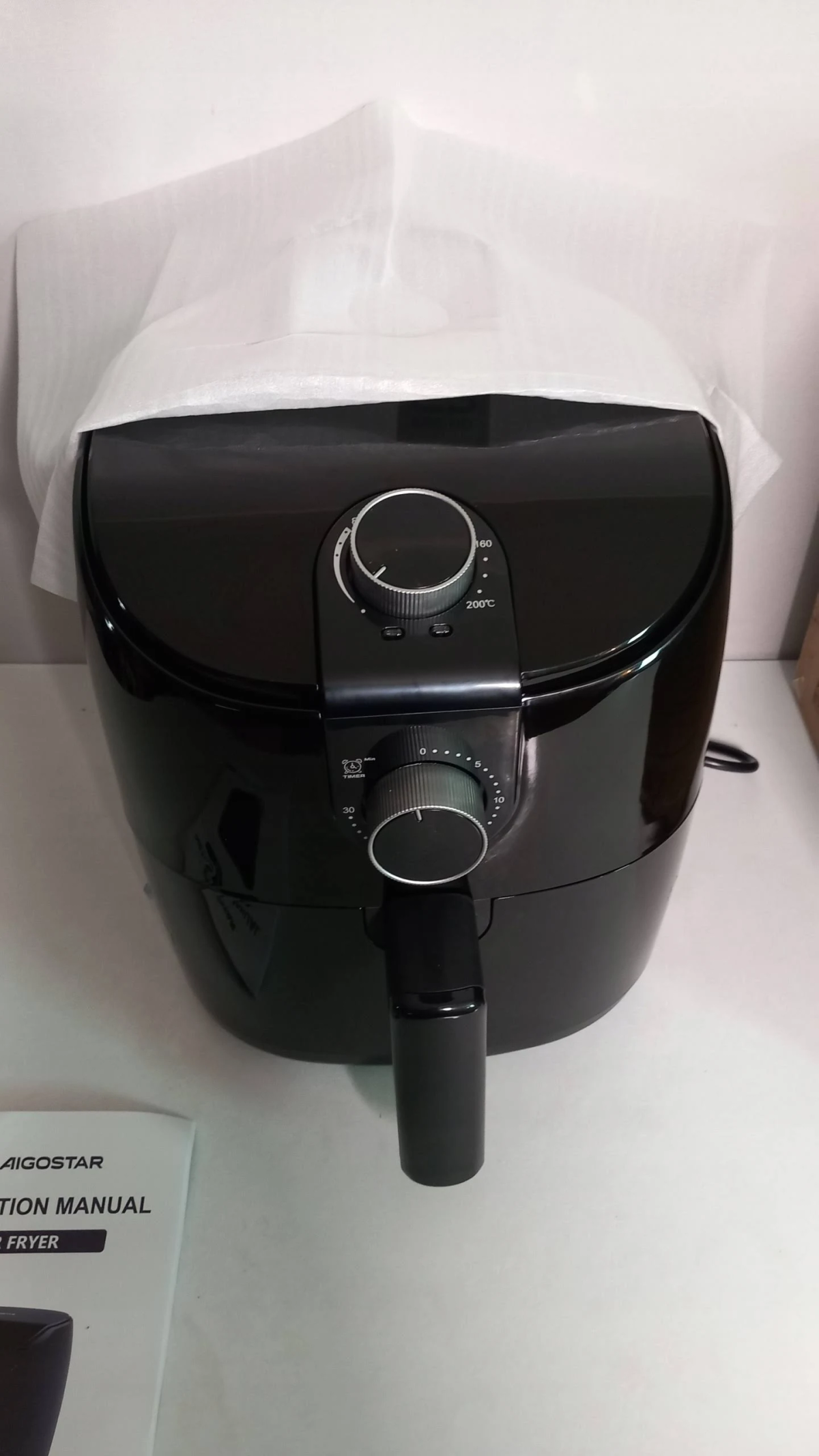 Schwarze Heißluftfritteuse in Verpackung mit Bedienungsanleitung. Marke ist AIGOSTAR.