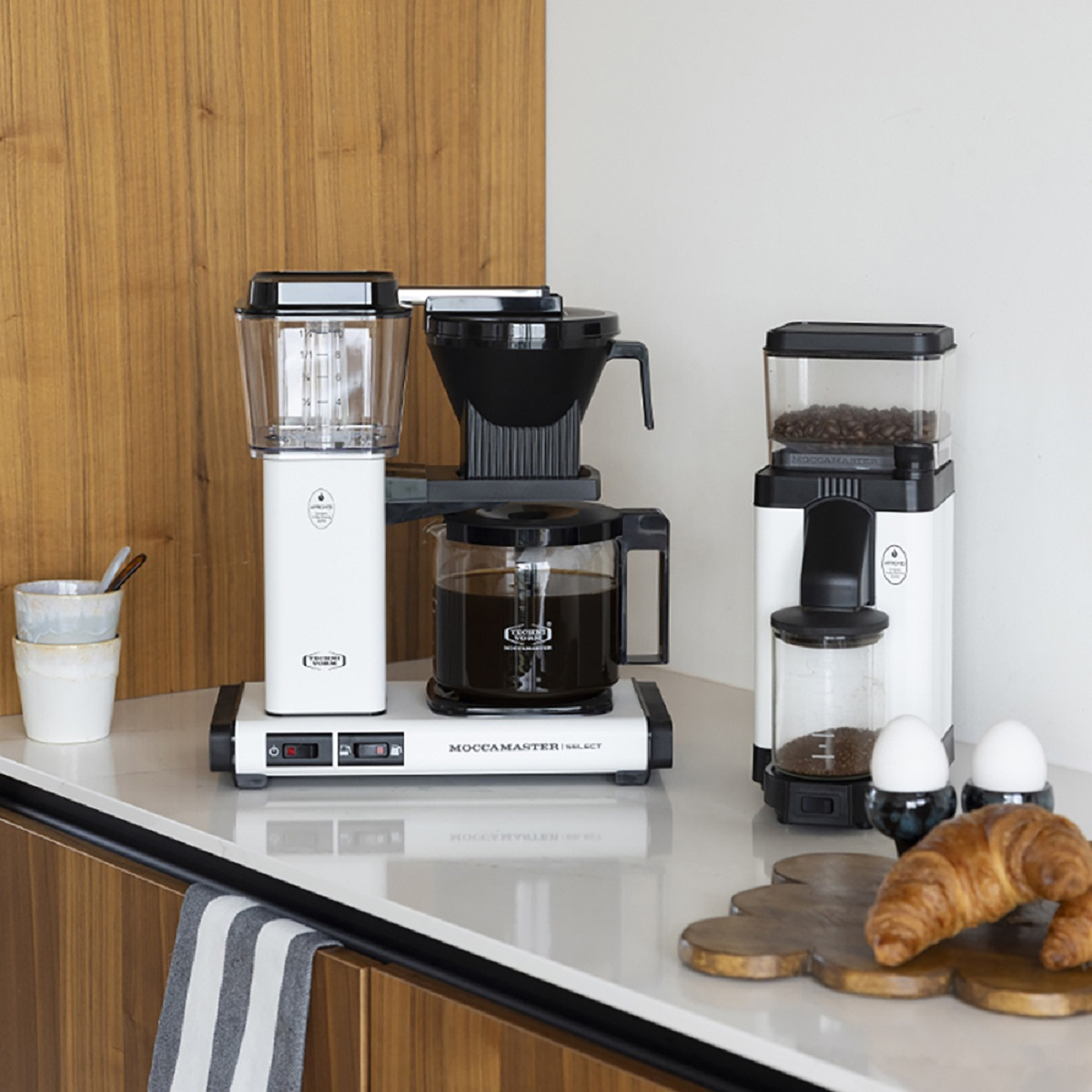 Ekspresy do kawy i młynek do kawy na blacie. Rogalik i jajka również na blacie.