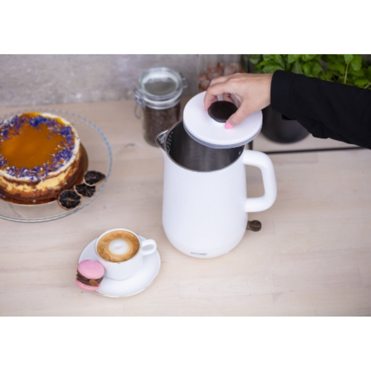 La mano di una donna che apre un bollitore elettrico bianco, con torta e caffè vicini.