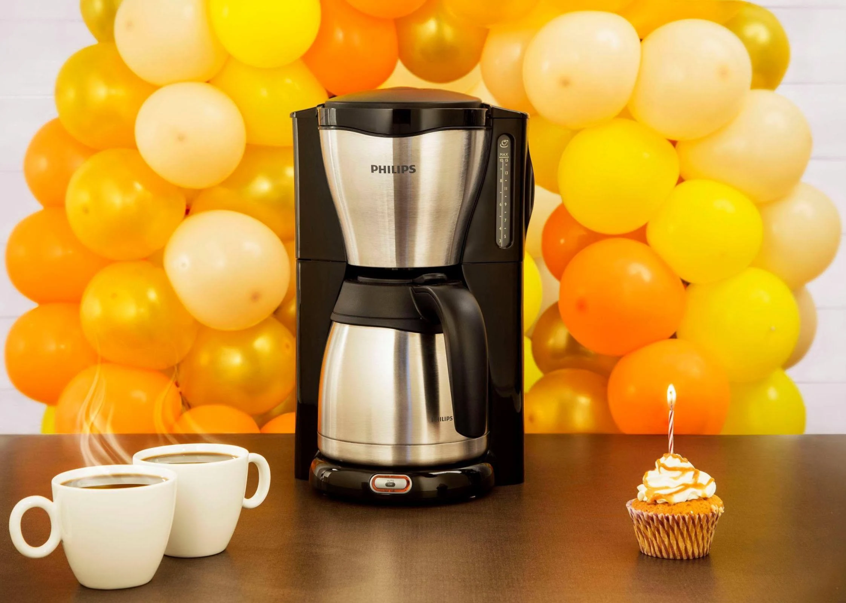 Caffettiera Philips nera e argento con due tazze bianche e un cupcake con candela.
