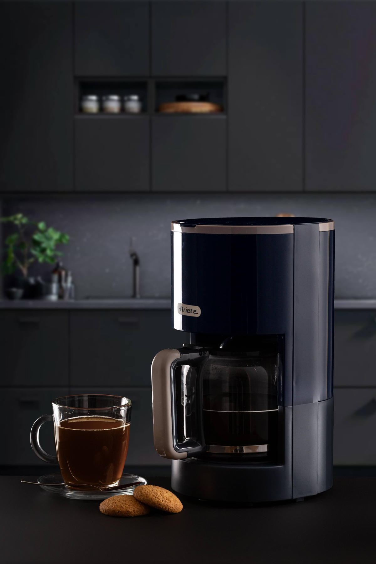 Ciemnoniebieski ekspres do kawy obok filiżanki kawy i ciasteczek.