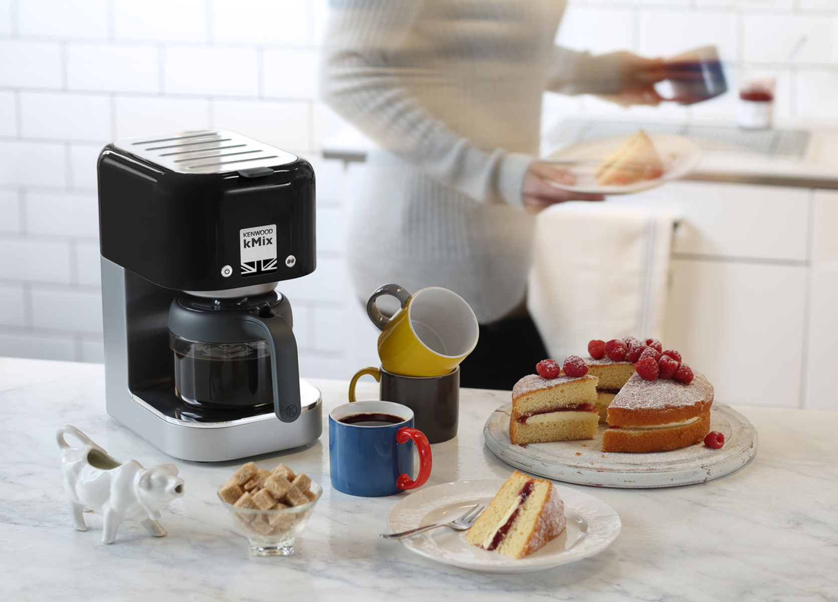 Scena di cucina con una caffettiera, torte e tazze. Una persona tiene dei piatti.