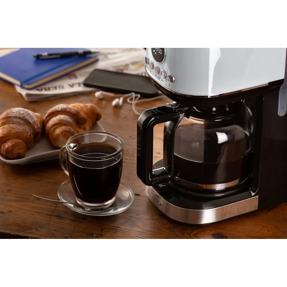 Ekspres do kawy ze szklaną karafką kawy, rogalikami i telefonem na drewnianym stole.