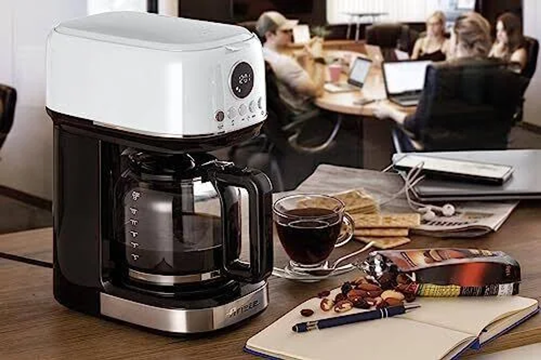 Czarno-biały ekspres do kawy na drewnianym stole. Filiżanka kawy, przekąski i ludzie w tle.