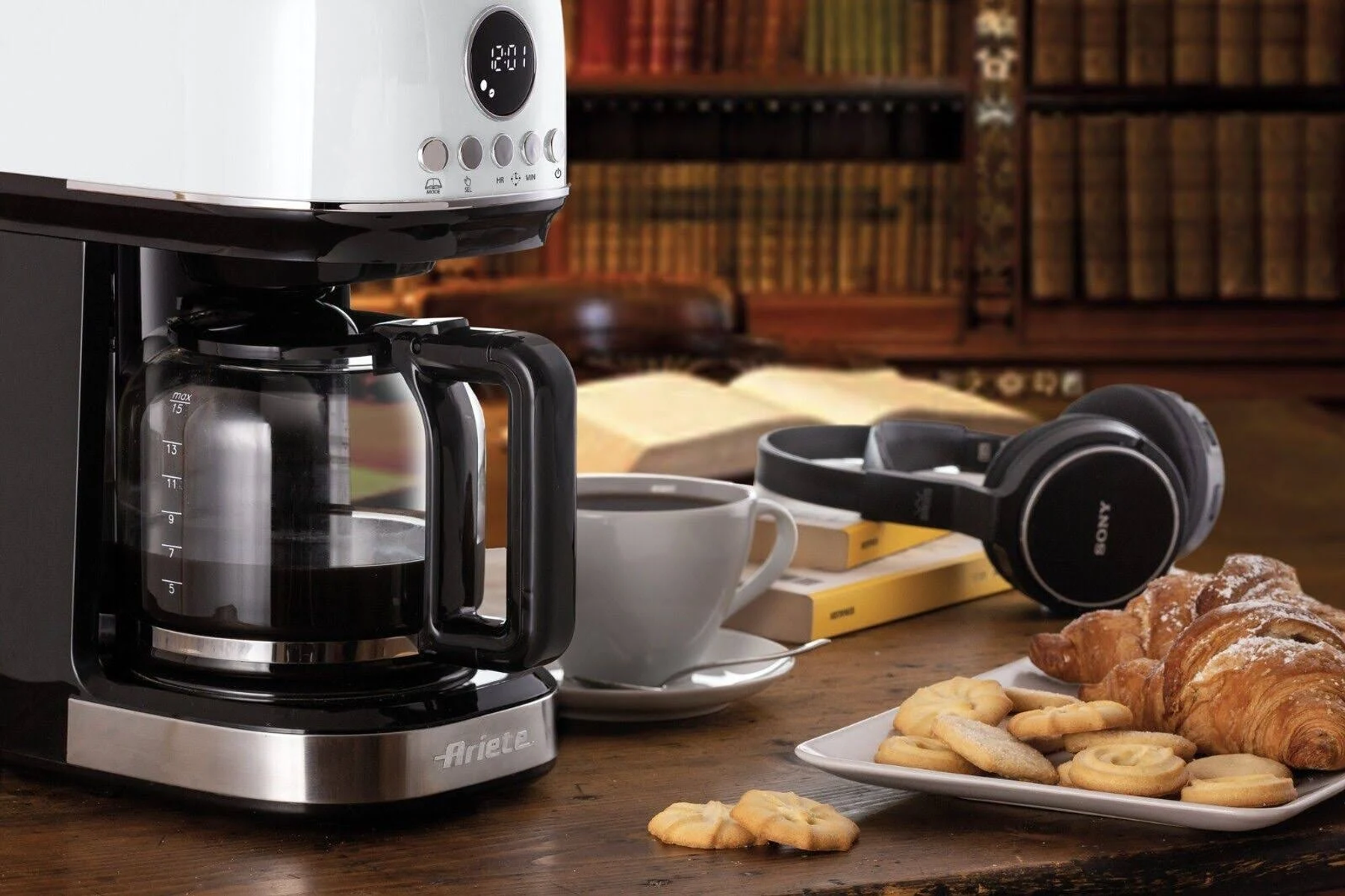 Ekspres do kawy na drewnianym stole z kawą, ciastkami, rogalikiem i słuchawkami. Książki w tle.