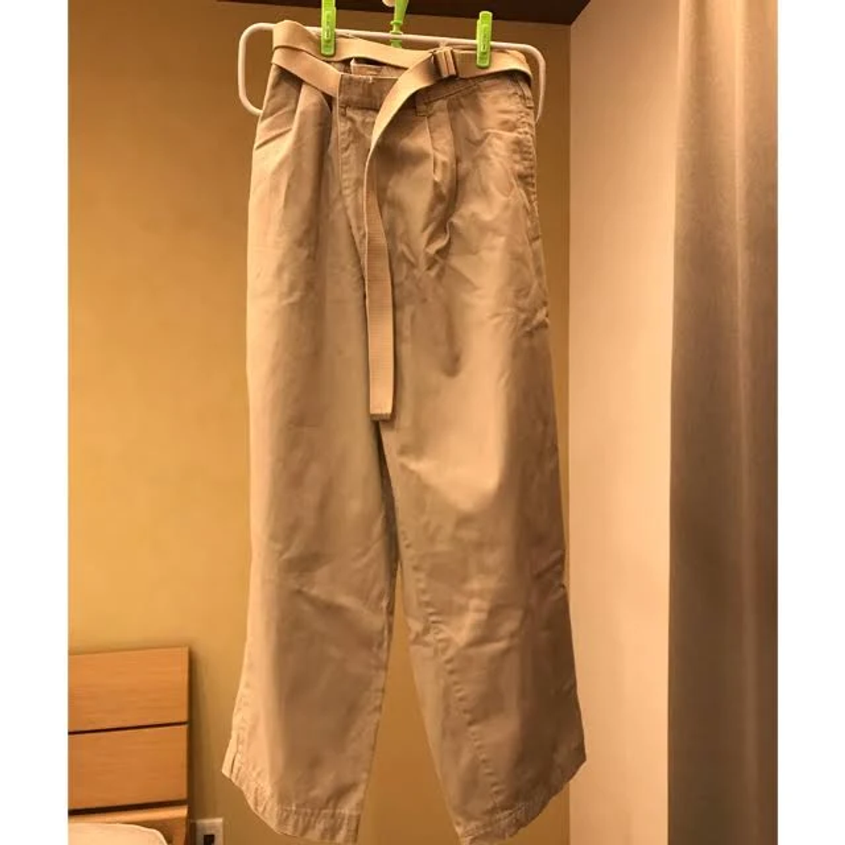 Beżowe spodnie khaki z paskiem wiszące na wieszaku. Jasne tło.