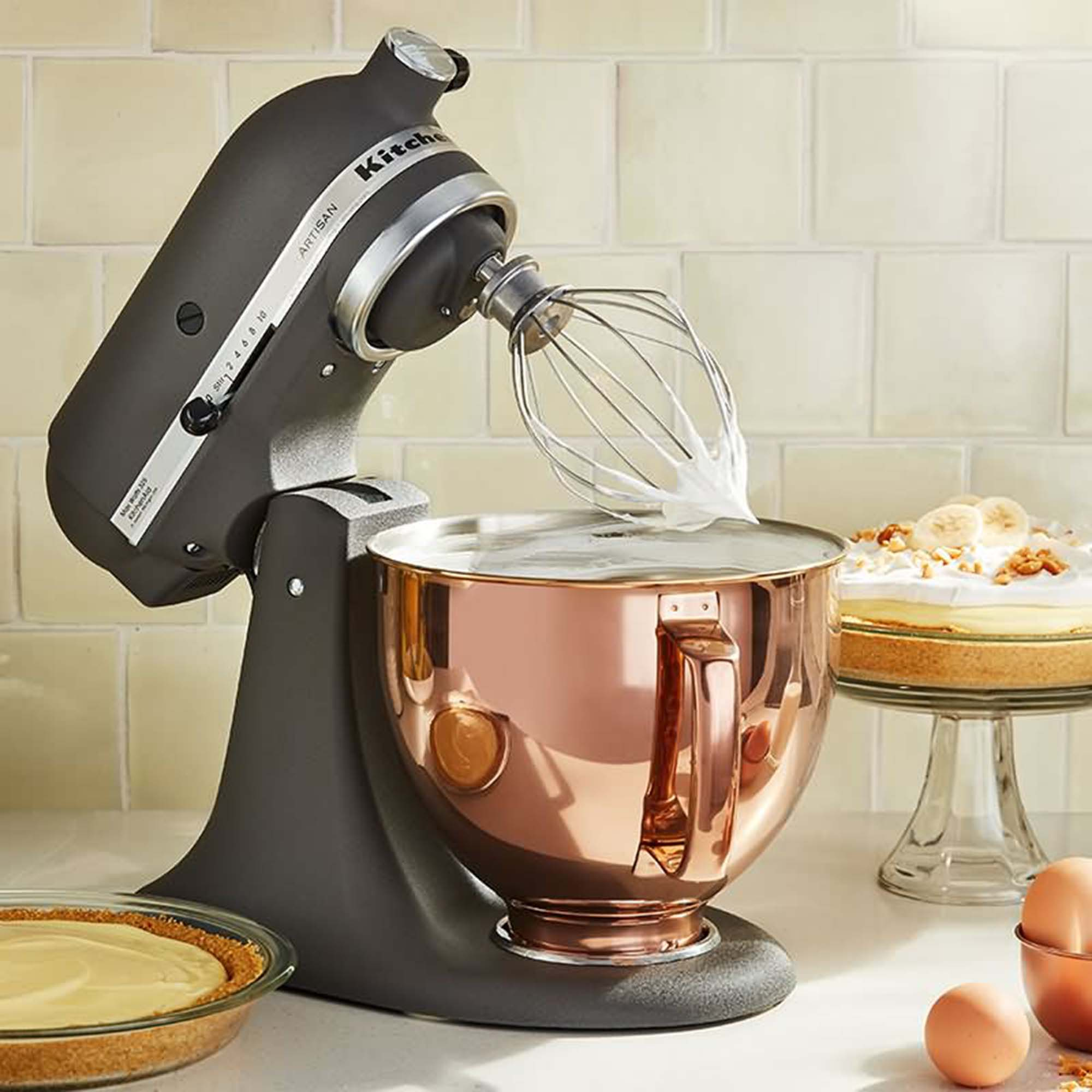 Ein grauer Standmixer mit einer Kupferschüssel, der Zutaten verrührt. Ein Kuchen und Käsekuchen sind an der Seite.