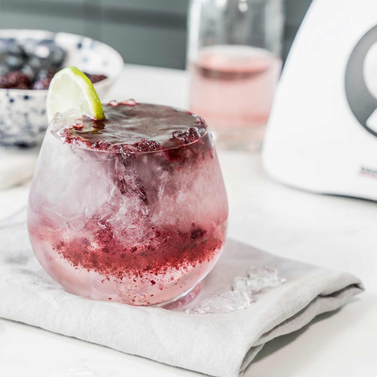 Ein Glas gemischtes Beerengetränk mit Eis, mit Limette garniert, auf einer grauen Serviette.