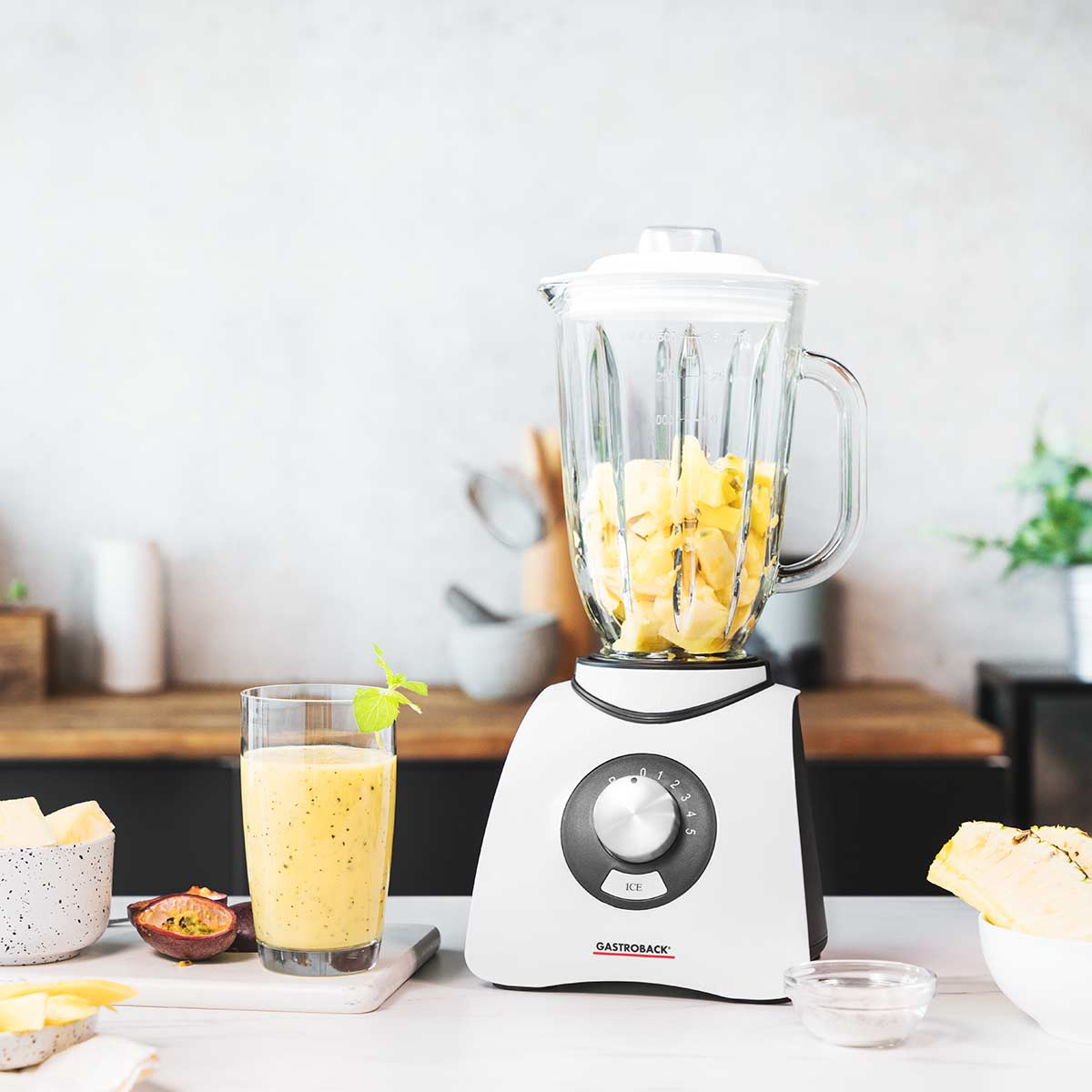 Mixer mit Zutaten auf einer weißen Arbeitsplatte. Ein Glas Smoothie und Obst.