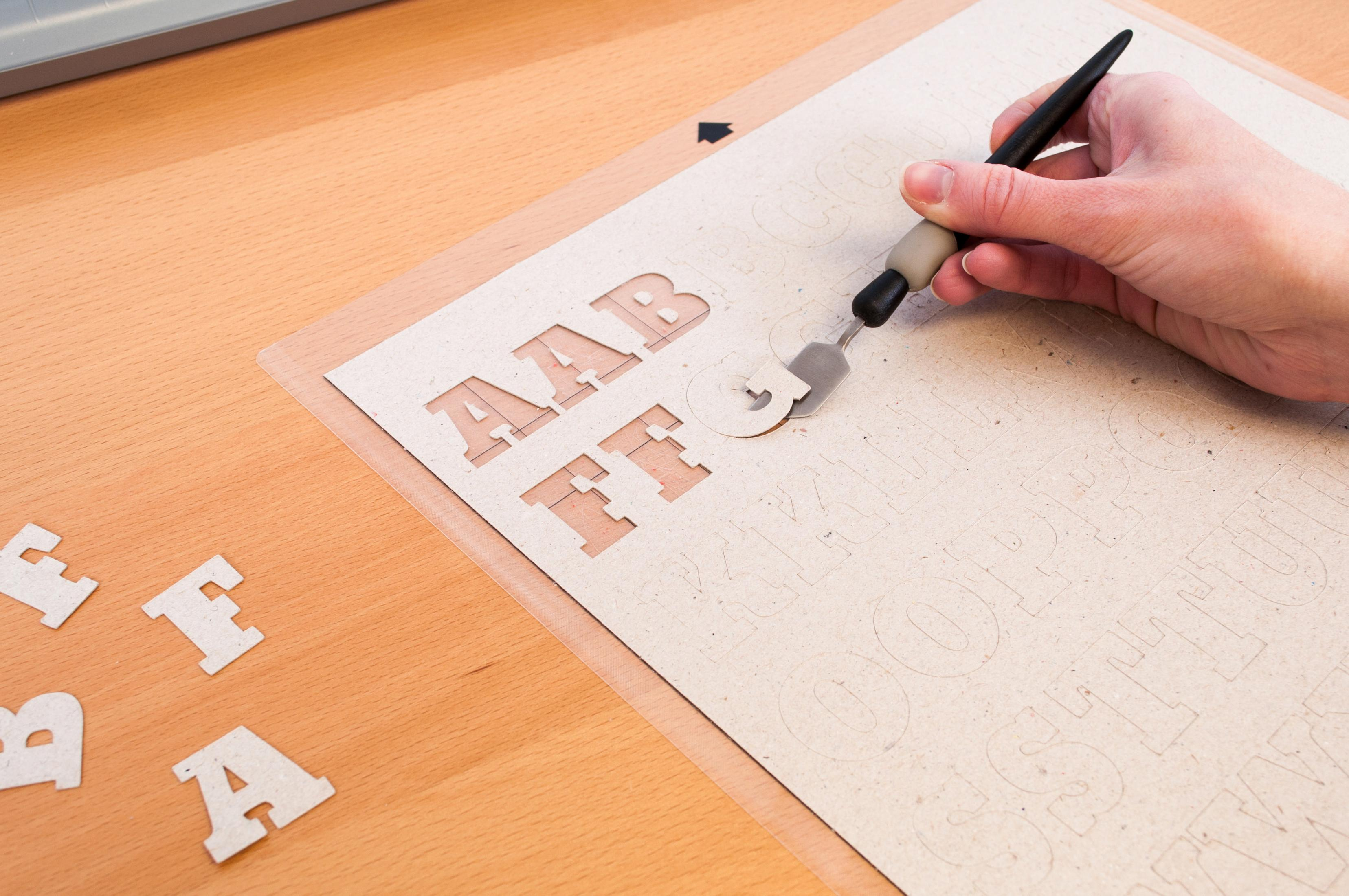 Eine Hand schneidet mit einem Bastelmesser Buchstaben aus einem Karton, ausgeschnittene Buchstaben liegen daneben.