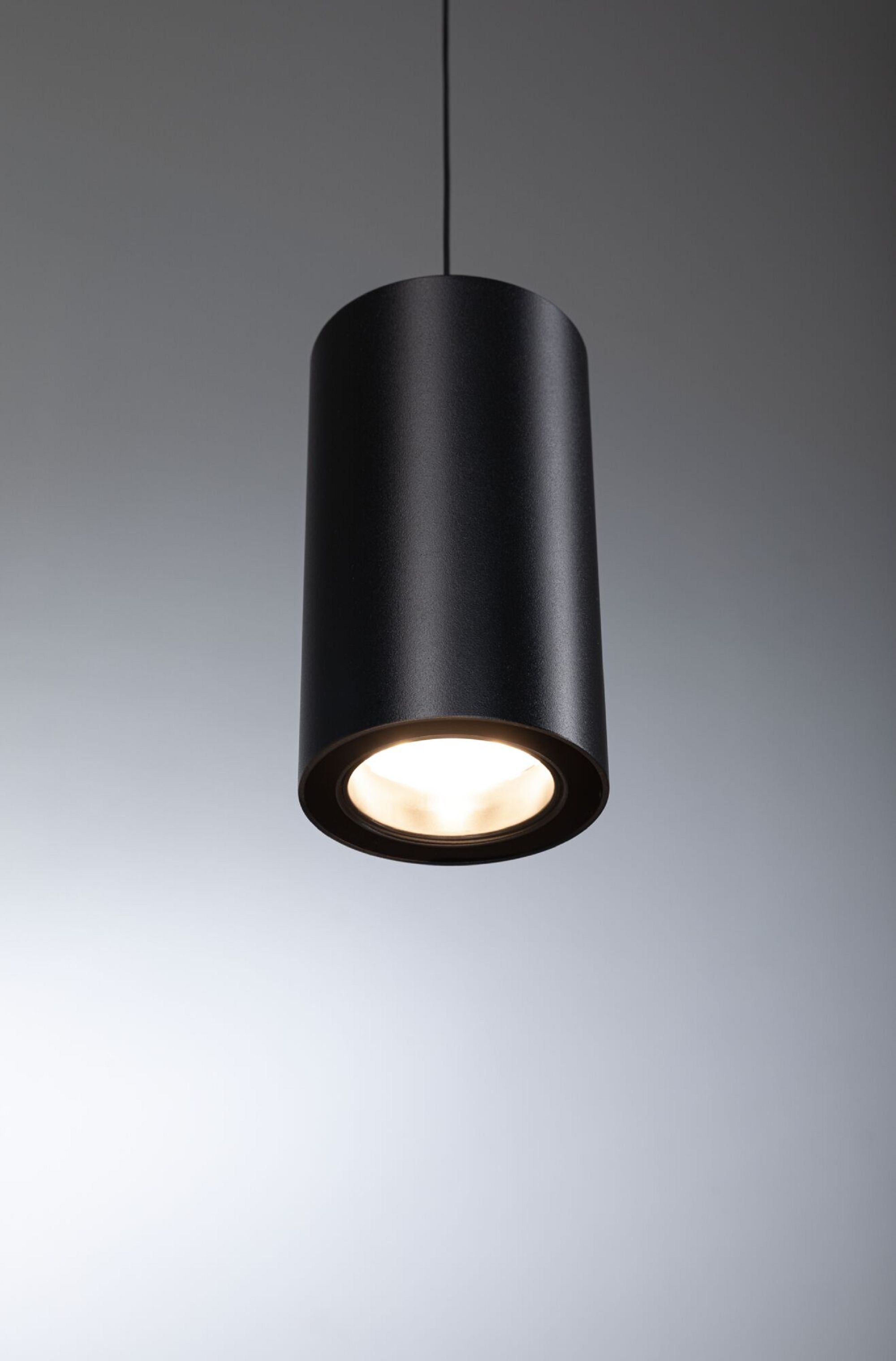 Schwarze zylindrische Pendelleuchte mit heller Lichtquelle. Von oben hängend, auf grauem Hintergrund.
