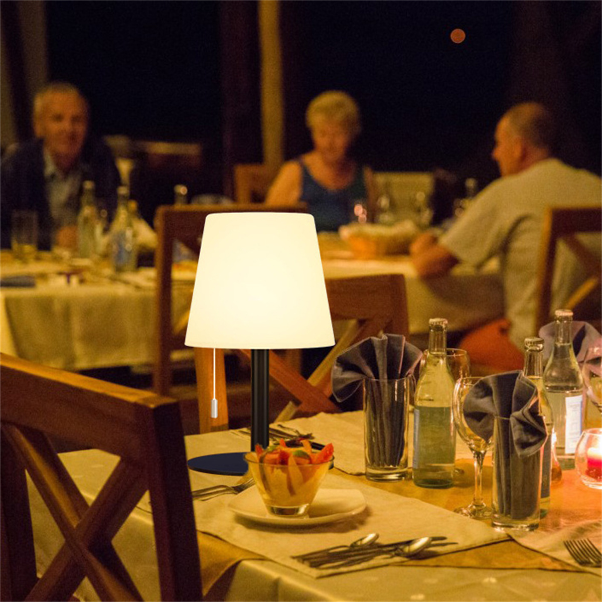 Tischlampe auf einem Tisch mit Essen und Weingläsern. Personen sitzen im verschwommenen Hintergrund.