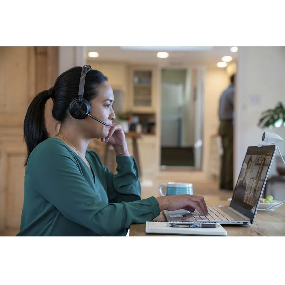 Mujer con auriculares trabajando en un portátil con una webcam en una cocina.