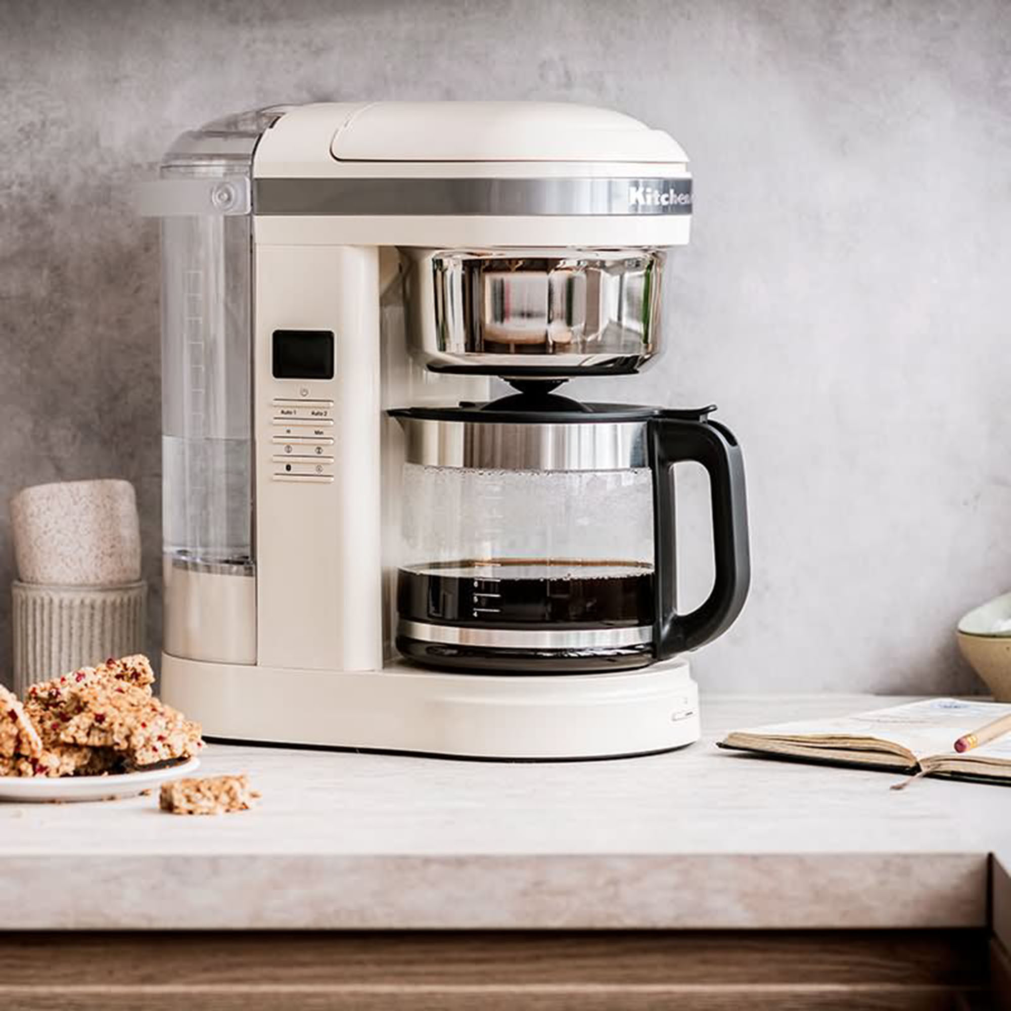 Beigefarbene KitchenAid Kaffeemaschine mit Glaskaraffe auf einer Arbeitsplatte mit Essen und Notizbuch.