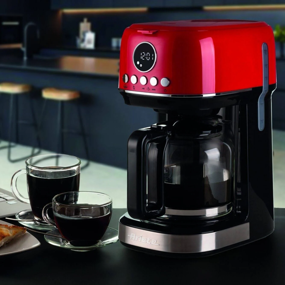 Una cafetera roja y negra con dos vasos de café sobre una mesa.