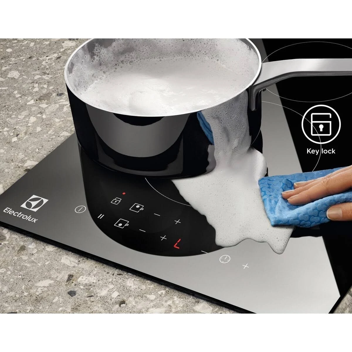 Un piano cottura a induzione nero con una pentola di acqua bollente e una mano che lo pulisce con un panno blu.