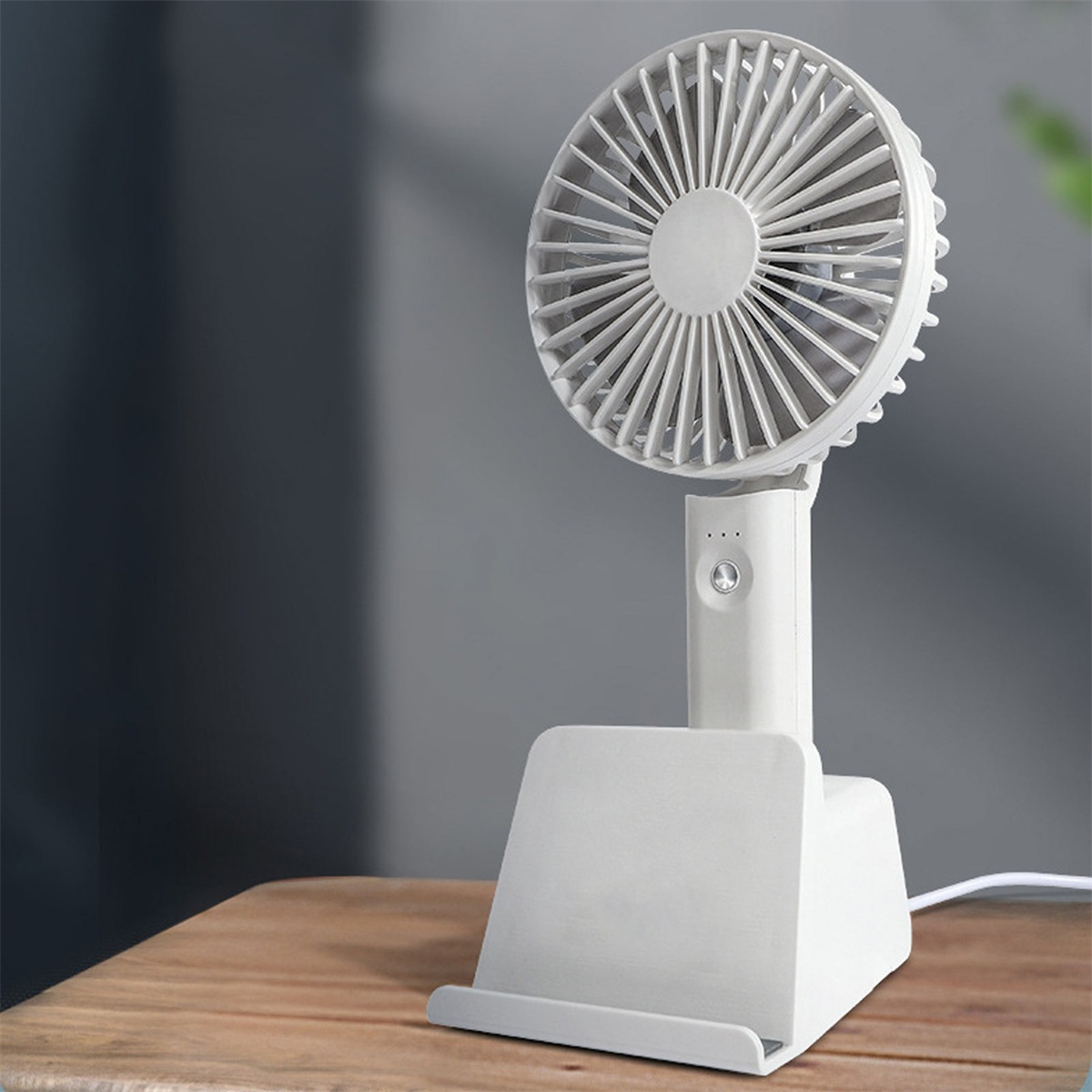 Ein weißer Tischventilator steht auf Holz. Der Ventilator hat einen Ständer. Der Hintergrund ist eine graue Wand.